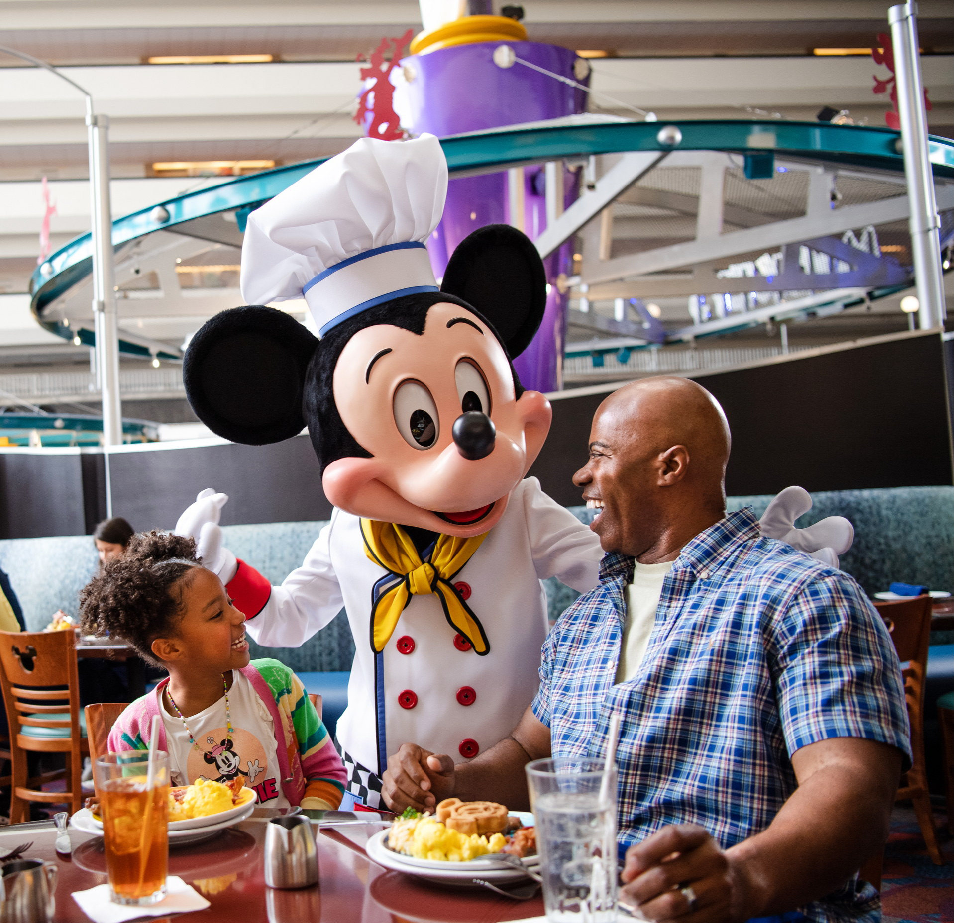 Mickey in a chef’s hat greets a girl and her father at their table beneath the monorail track, at Chef Mickey’s Mickey in a chef’s hat greets a girl and her father at their table beneath the monorail track, at Chef Mickey’s