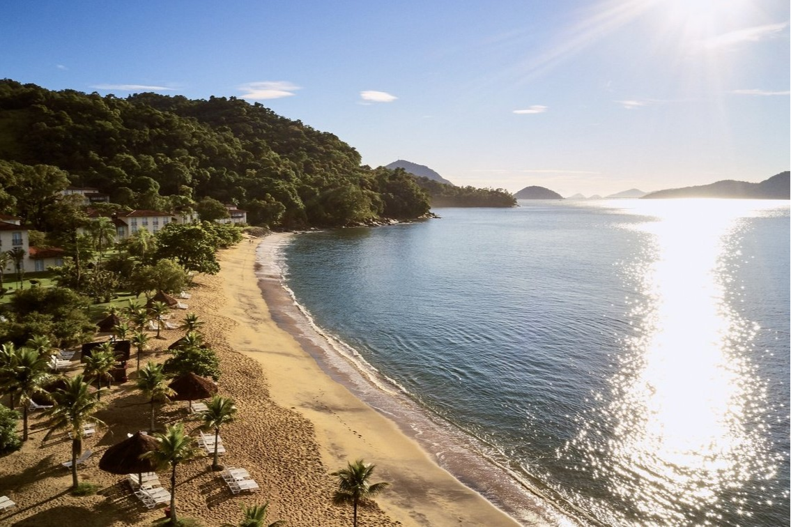 Aerial view of Club Med Rio Das Pedras with its palm-lined beach, lounge chairs, and a mountain backdrop. Aerial view of Club Med Rio Das Pedras with its palm-lined beach, lounge chairs, and a mountain backdrop.