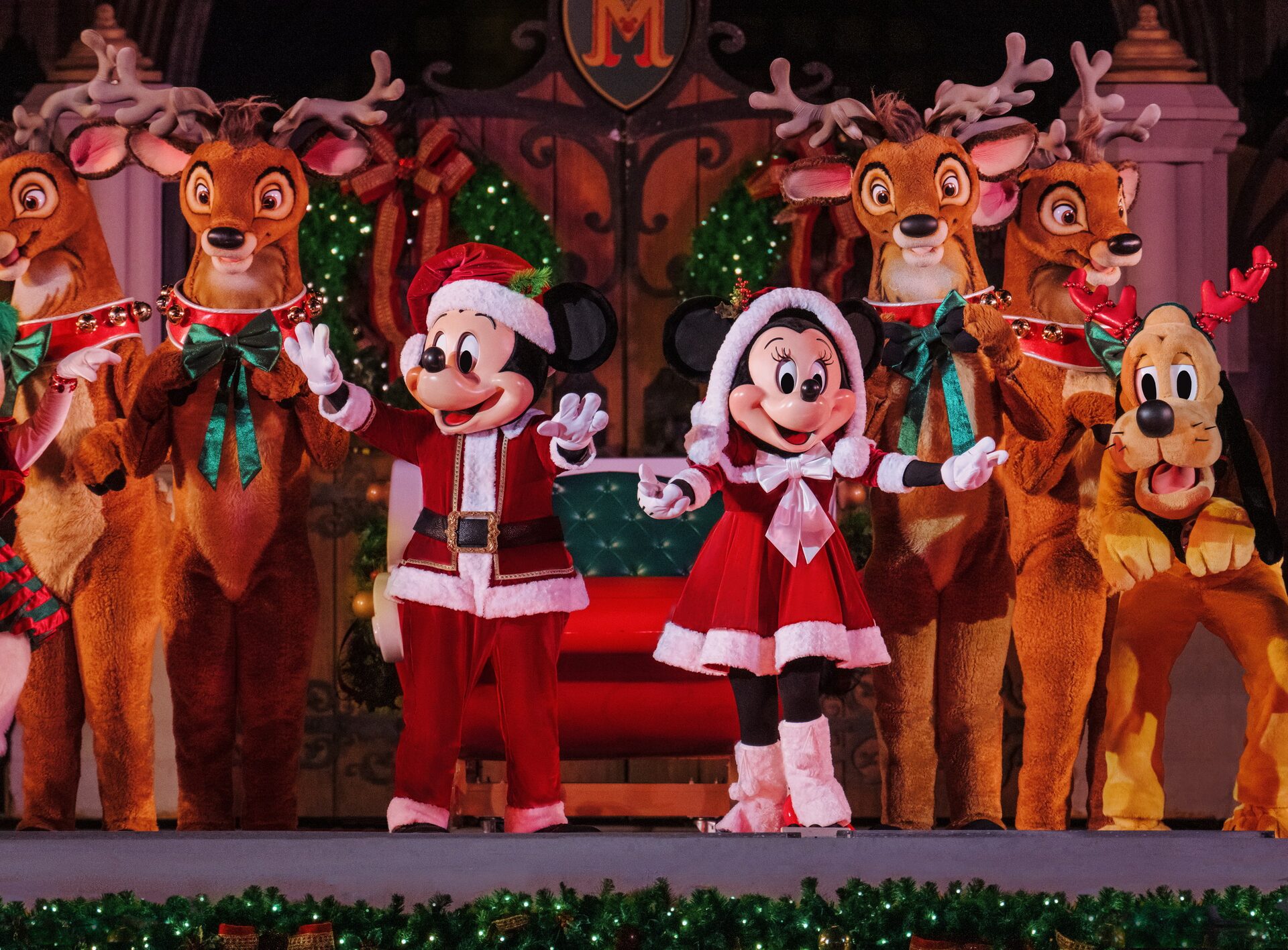 Mickey et Minnie en habits de père Noël saluent depuis un traîneau entouré de rennes costumés, sur une scène décorée de guirlandes, à Magic Kingdom Mickey et Minnie en habits de père Noël saluent depuis un traîneau entouré de rennes costumés, sur une scène décorée de guirlandes, à Magic Kingdom