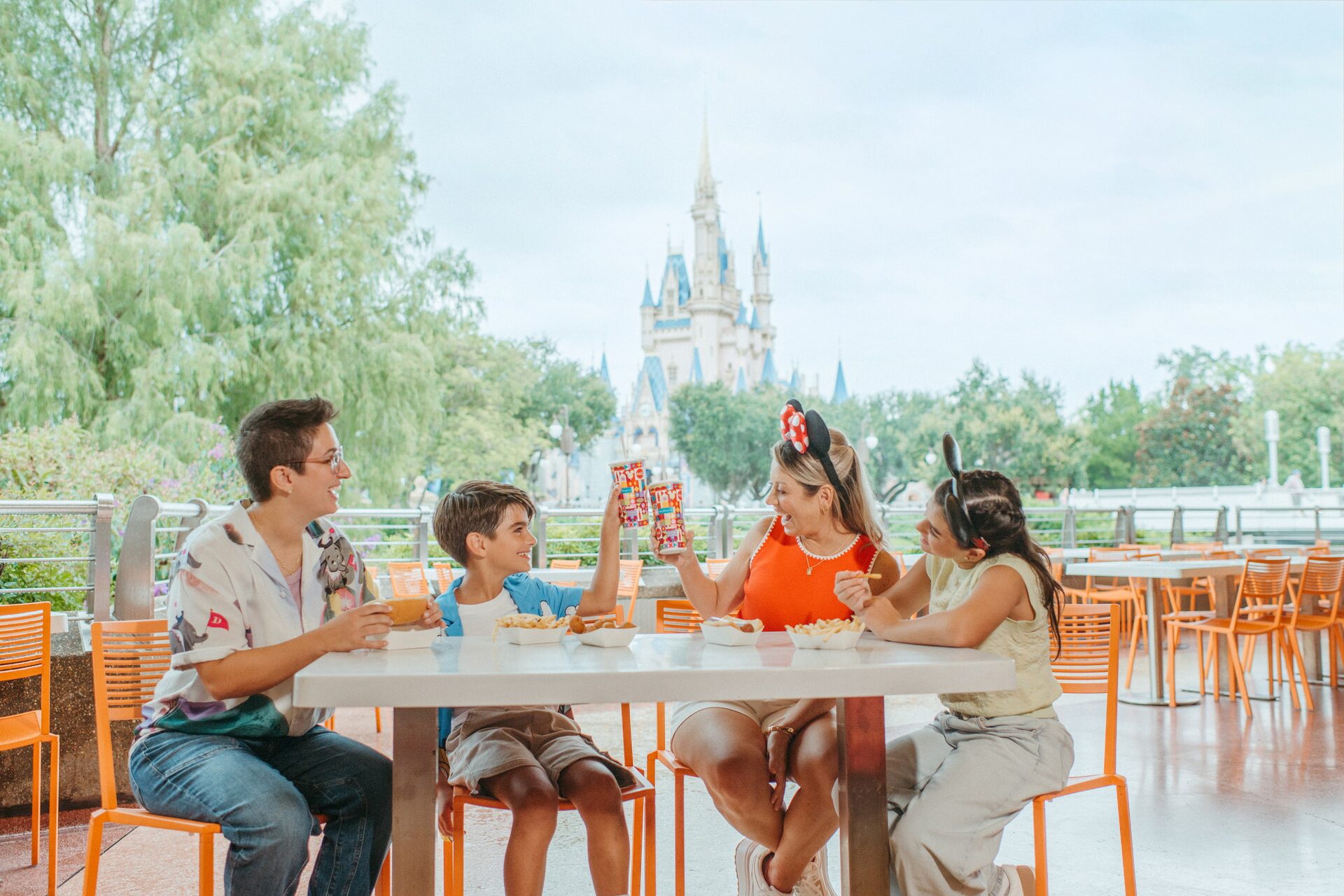 Quatre convives trinquent autour d’une table de terrasse orange, le château de Cendrillon au loin derrière les arbres, à Magic Kingdom Quatre convives trinquent autour d’une table de terrasse orange, le château de Cendrillon au loin derrière les arbres, à Magic Kingdom