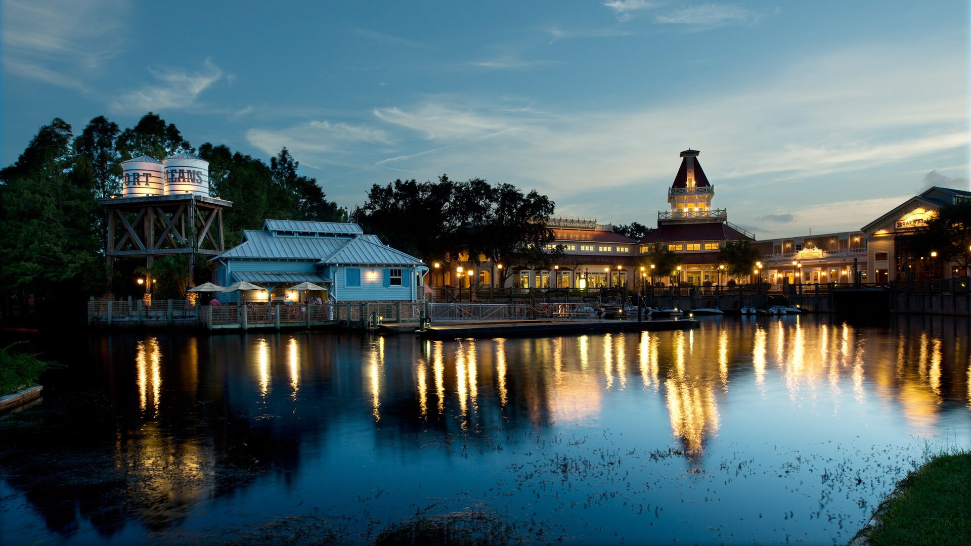 Les bâtiments éclairés du Disney’s Port Orleans Resort – Riverside se reflètent dans la rivière au crépuscule, le château d’eau à gauche Les bâtiments éclairés du Disney’s Port Orleans Resort – Riverside se reflètent dans la rivière au crépuscule, le château d’eau à gauche